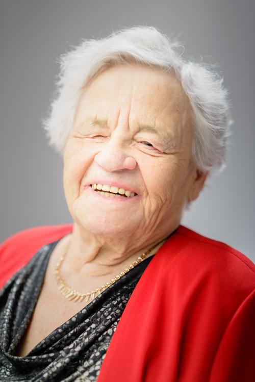 Muotokuva 84-vuotiaasta naisesta. Hän hymyilee niin että hampaat näkyvät. Hänellä on harmaat, lyhyet, tuuheat ja hieman kihartuvat hiukset. Hänellä on kultainen kaulakoru. Yllään hänellä on pusero, jossa on mustalla pohjalla harmaata printtikuvioita, joka kuvaa koristeellisia puun oksia ja lehtiä. Puseron päällä on kirkkaanpunainen jakku. 